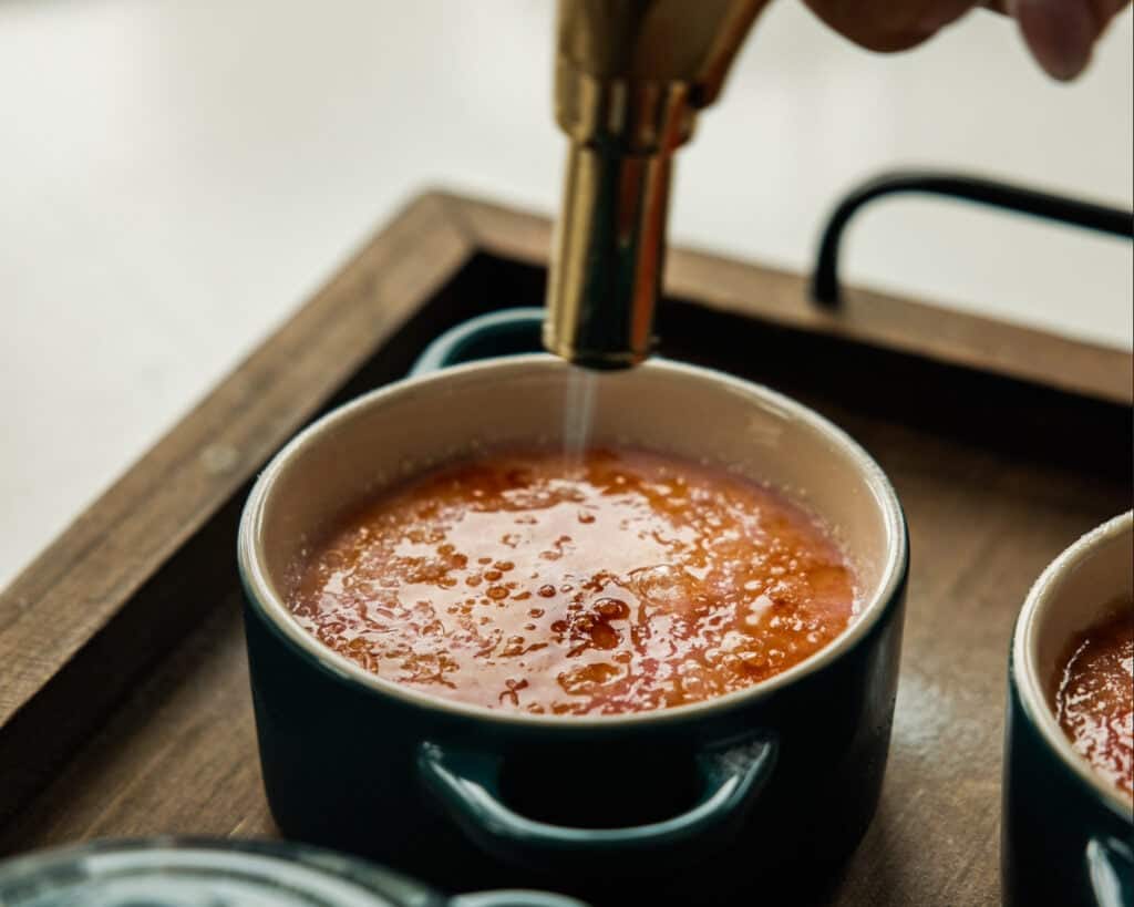 A close-up of a hand using a torch to caramelize the sugar on top of an Easy Crème Brûlée in a small green ramekin on a wooden tray.